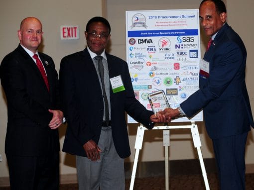 Three men are at a podium with a sign displaying sponsors for the 2018 Procurement Summit. Two of the men are exchanging a trophy, while the third stands beside them, smiling.