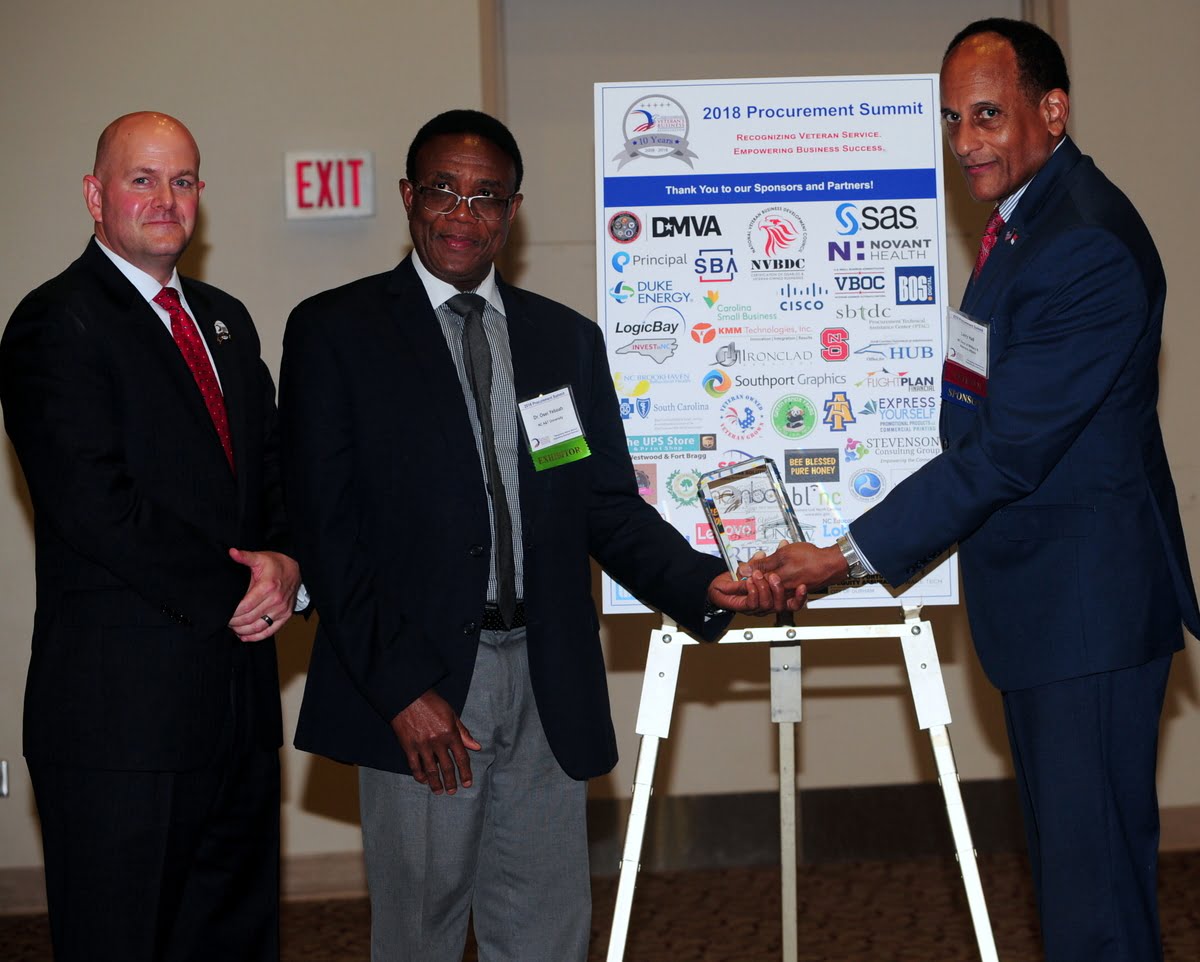 Three men are at a podium with a sign displaying sponsors for the 2018 Procurement Summit. Two of the men are exchanging a trophy, while the third stands beside them, smiling.