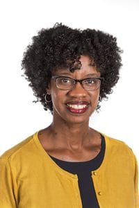 A woman with curly black hair and glasses smiles, wearing a mustard-colored cardigan over a black shirt.