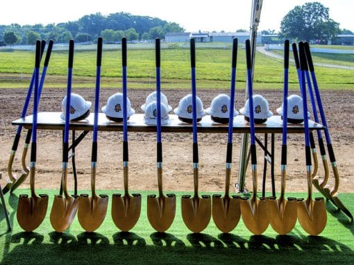 A table displays multiple gold shovels and white hard hats with a logo, set against a grassy field and blue sky.