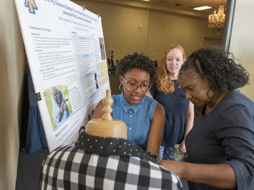 Three people engage around a mannequin display in front of a research poster. One woman with glasses and a denim shirt talks while another woman observes, and a third examines the garment on the mannequin. Background elements include a chandelier and a table.