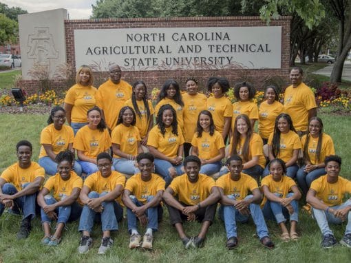 A diverse group of students wearing matching yellow shirts poses in front of a sign for North Carolina Agricultural and Technical State University. They sit on the grass, surrounded by flowers, and display a sense of unity and camaraderie.