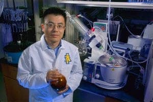 A scientist in a laboratory wears a white lab coat with a logo. He holds a brown flask and stands near scientific equipment, including a distillation apparatus and other glassware on a lab countertop. The setting is well-lit with various lab tools in the background.
