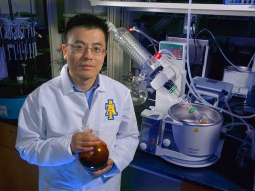 A scientist in a laboratory wears a white lab coat with a logo. He holds a brown flask and stands near scientific equipment, including a distillation apparatus and other glassware on a lab countertop. The setting is well-lit with various lab tools in the background.