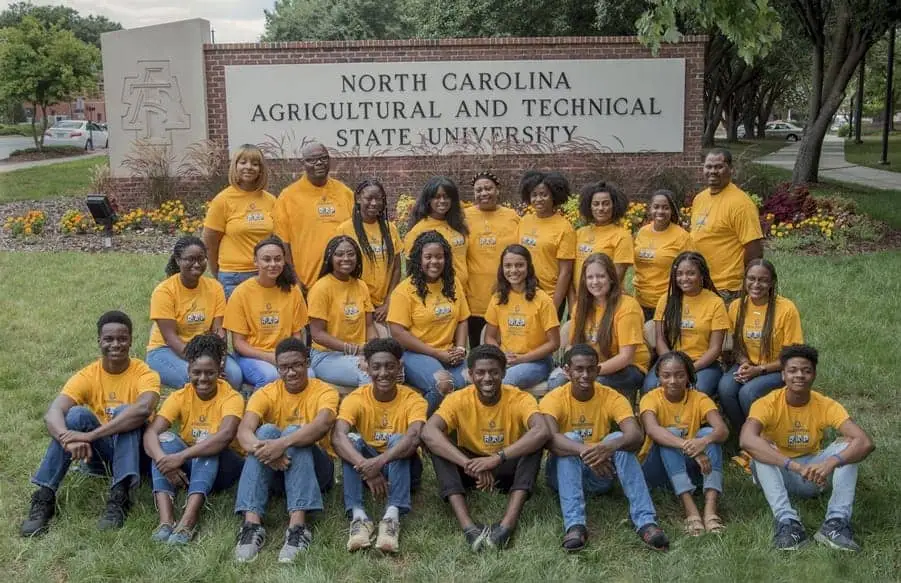 A diverse group of students wearing matching yellow shirts poses in front of a sign for North Carolina Agricultural and Technical State University. They sit on the grass, surrounded by flowers, and display a sense of unity and camaraderie.