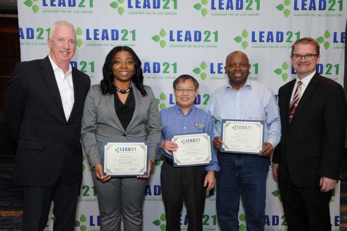 Five people stand together, holding certificates, in front of a backdrop for LEAD21. The group includes three men and two women, all dressed in professional attire.