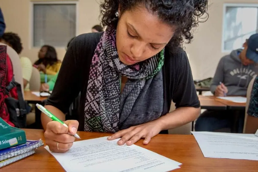 Woman studying and writing notes in a classroom setting with other students.