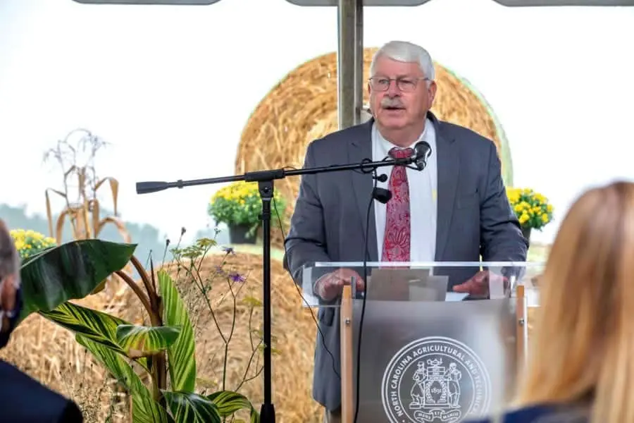 Agriculture official speaking at outdoor event with hay bales and plants in background.
