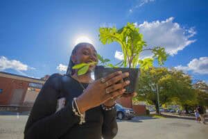 A woman smiles while holding a potted plant against a bright blue sky with fluffy clouds.