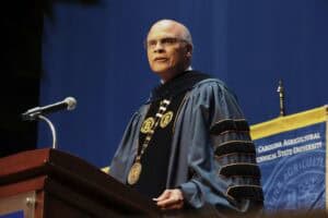 A speaker in academic regalia delivers a speech at a university event, with a podium and university banner behind him.