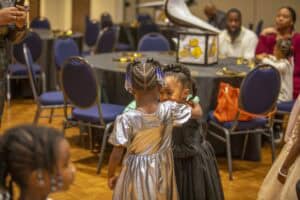 Two young girls share a warm embrace, dressed in silver and black dresses, at a decorated event venue.