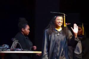 A smiling graduate in a cap and gown waves, while another attendee prepares behind her.
