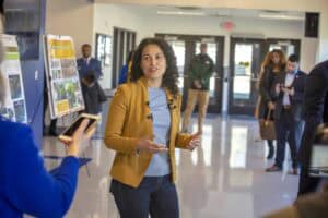 A woman in a mustard jacket gestures while engaged in conversation, with people and displays in the background.