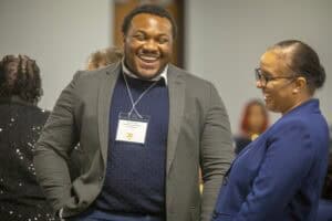 Two smiling people engage in conversation at an event, one wearing a blazer with a name tag.