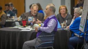 A group of people engaged in discussion at a round table, with one man in a purple sweater sitting thoughtfully.