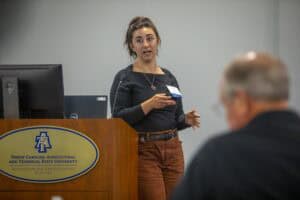 A woman speaks at a podium with a computer, engaged in a presentation while attendees listen.