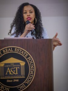 A woman with curly hair speaks passionately into a microphone at a podium featuring a university logo.