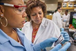 Dr. Mulumebet Worku, in a lab coat, working closely with a student in a laboratory setting at N.C. A&T. The student, also in lab attire and wearing protective goggles, holds a slide as Dr. Worku observes. In the background, other lab members work at their stations, indicating a busy research environment.