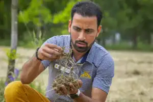 Soil scientist Dr. Biswanath Dari closely examining a clump of soil and roots outdoors. He wears a striped polo shirt with the North Carolina A&T logo, appearing focused as he inspects the sample. The background includes greenery, suggesting a natural or field setting.