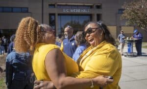 Two women in yellow tops embrace and laugh joyfully outside B.C. Webb Hall, surrounded by a lively crowd.