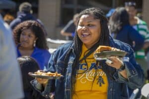 A woman in a denim jacket smiles as she carries two plates of food at an outdoor gathering.