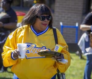 A woman in a yellow jacket holds a plate of food and a drink at an outdoor event, smiling in the sunlight.