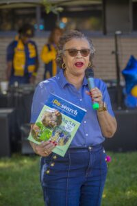 A woman speaks into a microphone while holding a magazine titled "Re:search Ag 5.0," wearing sunglasses and a blue…