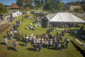Aerial view of an outdoor community gathering on a sunny day, featuring a large white tent with people seated underneath and a food serving area with people in line. Attendees are spread across the grassy area, with a food truck visible on the left and parking lot and buildings in the background