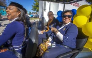 Three women in navy outfits smile and enjoy a festive outdoor event, surrounded by yellow and blue balloons.