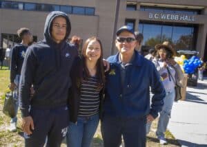Young man, woman, and older man posing together outside a building, smiling. A crowd and decorations are in the…