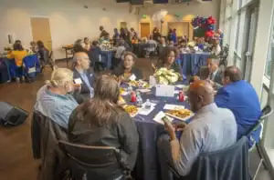People seated around a round table enjoying a meal at a formal event with multiple tables set up in a well-lit room. Attendees are engaged in conversation, with flower centerpieces and plates of food on the tables. The background shows other groups seated at tables, along with colorful balloon decorations and open windows letting in natural light.