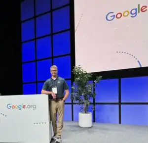 Man standing on stage beside a podium with the Google.org logo, smiling at the camera. Behind him, a large screen displays the Google logo, and a potted plant decorates the stage. The backdrop features a blue and black grid pattern, creating a modern, professional setting.