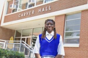 Jaliyah Shepherd, a Landscape Architecture Foundation Ignite Scholar, standing outside Carver Hall with a confident smile. She wears a white shirt under a blue sweater vest, with her hands relaxed in front of her. The brick building behind her displays the name 'Carver Hall' above the entrance."