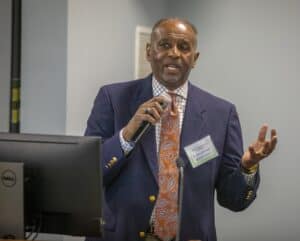 A man in a suit speaks at a conference, holding a microphone and gesturing. A computer screen is visible nearby.