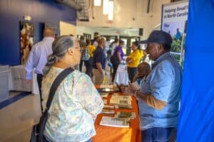 A woman speaks with a man at a community event, surrounded by informational tables and attendees.