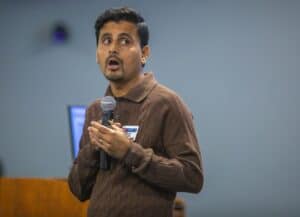 A man in a brown sweater speaks into a microphone, engaged in a presentation.