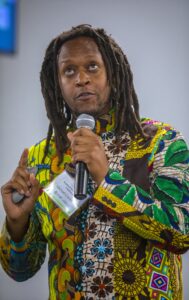 Empowered speaker with dreadlocks wearing vibrant African print shirt, holding a microphone at a con.