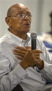 An older man with glasses speaks passionately into a microphone, hand over his heart, in a casual setting.