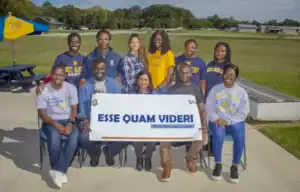 Group photo of the team Esse Quam Videri, which translates to 'To be rather than to seem,' holding a banner with their team name. The group of eleven people smiles at the camera, with a grassy field and farm buildings in the background. Some team members wear North Carolina A&T State University apparel.