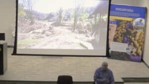 Presenter stands in front of a large projector screen displaying a landscape affected by environmental damage, with cleared trees and dirt paths. To the side, a banner for the College of Agriculture and Environmental Sciences highlights images related to research and nutrition. The presenter gestures towards the audience in a lecture hall.