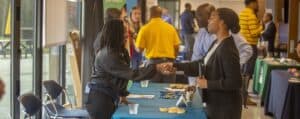 Two individuals shake hands across a table at a career fair, with brochures, pens, and snacks displayed on the table. Other attendees and booths are visible in the background.
