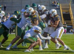 Players from North Carolina A&T and an opposing team clash during a football game. A North Carolina A&T player, wearing a black jersey and white helmet, carries the ball while teammates block defenders dressed in white and green jerseys.