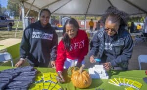 Three women smiling and interacting at a promotional table under a tent. The table is decorated with pumpkins and covered with branded items, including Syngenta notebooks, beanies, and pens. Two women wear Syngenta-branded hoodies, and one wears a red hoodie labeled "WFEY."
