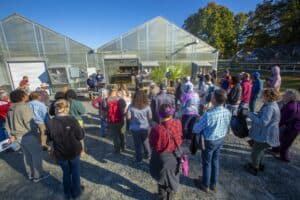 A group of attendees gathers in front of two large greenhouses during a field day event. Two speakers stand at a table, presenting information. Potted plants and equipment are displayed, and the event takes place under a clear blue sky.