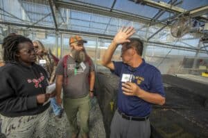 Guochen Yang, Ph.D., wearing an NC A&T polo, speaks to a group of farmers inside a greenhouse during a field day event. He gestures with one hand while explaining, as attendees listen attentively. The greenhouse structure, with fans and agricultural equipment, is visible in the background.
