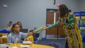 Michael Carter Jr., wearing a colorful patterned shirt, hands a green baton to a seated participant during a demonstration of "passing the baton" at a conference session. The participant, dressed in a blue button-down shirt, reaches out to accept the baton while other attendees watch in the background. The scene takes place in a conference room with yellow tablecloths and informational banners visible in the background.
