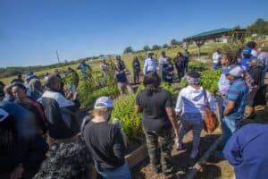 Hannah Talton stands in the center of a community garden, gesturing as she directs a presentation to a group of participants. The group forms a semi-circle around her, with individuals listening attentively, taking photos, and holding materials. The garden features raised beds of plants, and the background shows an open field, a wooden pavilion, and a clear blue sky.