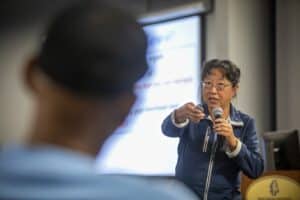 Kathleen Liang, Ph.D., speaks into a microphone and gestures with a presentation remote while addressing an audience during a presentation. A blurred participant's head is visible in the foreground, and a screen with text is in the background.