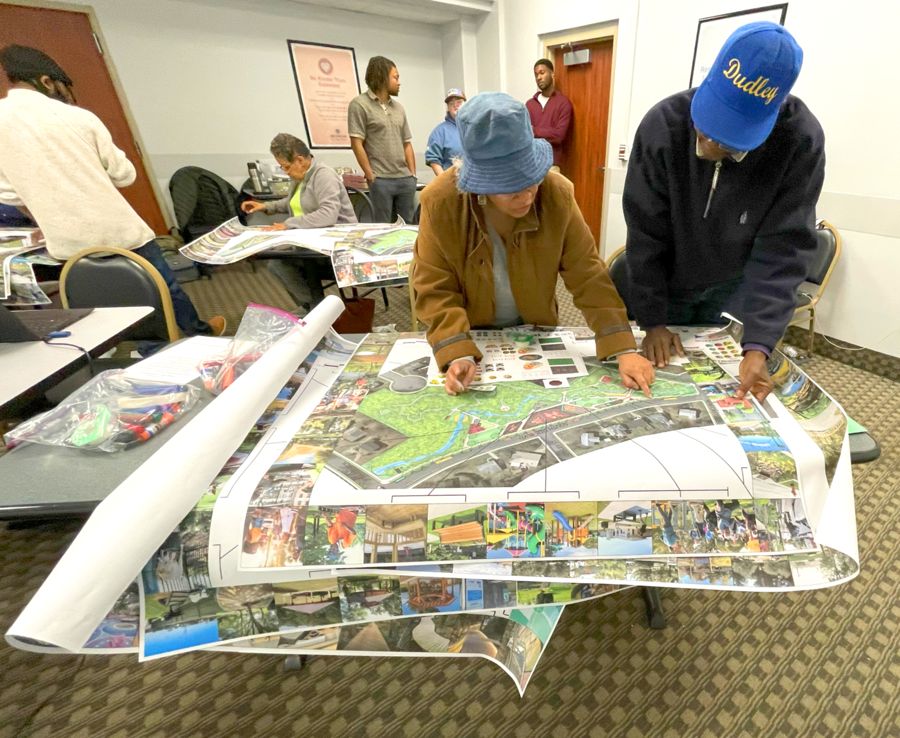 A group of people in a community meeting setting are gathered around tables, reviewing large maps and plans for a park or public space. Two individuals in the foreground are leaning over a map, pointing and discussing details. One is wearing a blue bucket hat, and the other is wearing a blue cap that reads "Dudley." Others in the background are seated or standing, engaged in similar activities. The room is bright with white walls, and materials like markers and papers are visible on the tables.