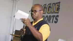 A middle-aged man with glasses and a beard, wearing a yellow polo shirt and a navy blue vest, speaks into a microphone while holding a piece of paper. He appears to be delivering a speech or presentation. Behind him, a wall displays the words 